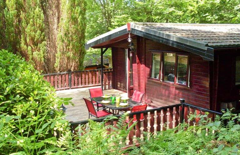 A wooden cabin with a deck and red chairs surrounded by green trees and plants at Top Lodge 4 Skiptory Howe White Cross Bay near Troutbeck Bridge