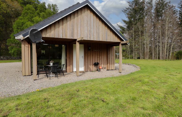 A wooden cottage with an outdoor table and chairs on gravel next to a grassy lawn and trees at Stone Water Cottage in Strathpeffer