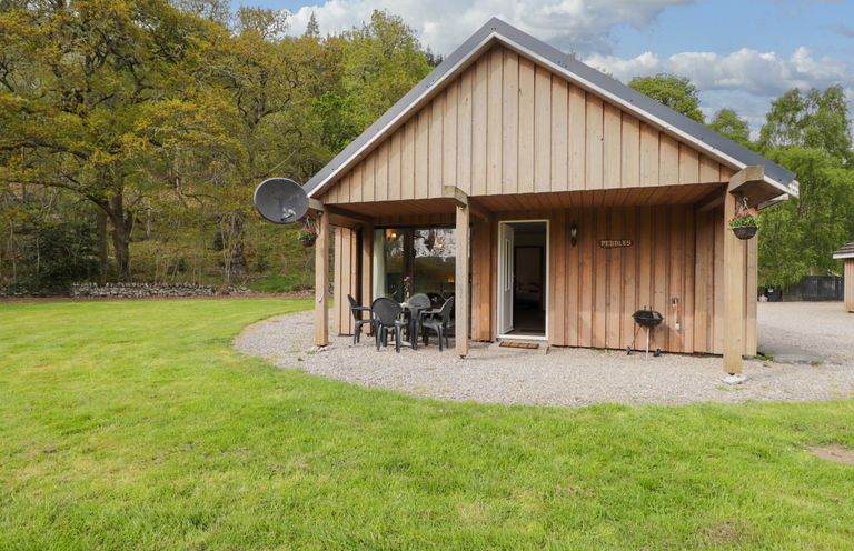 The exterior of a wooden cottage with a covered porch table and chairs on a gravel area surrounded by grass at Pebbles Cottage in Achilty near Strathpeffer
