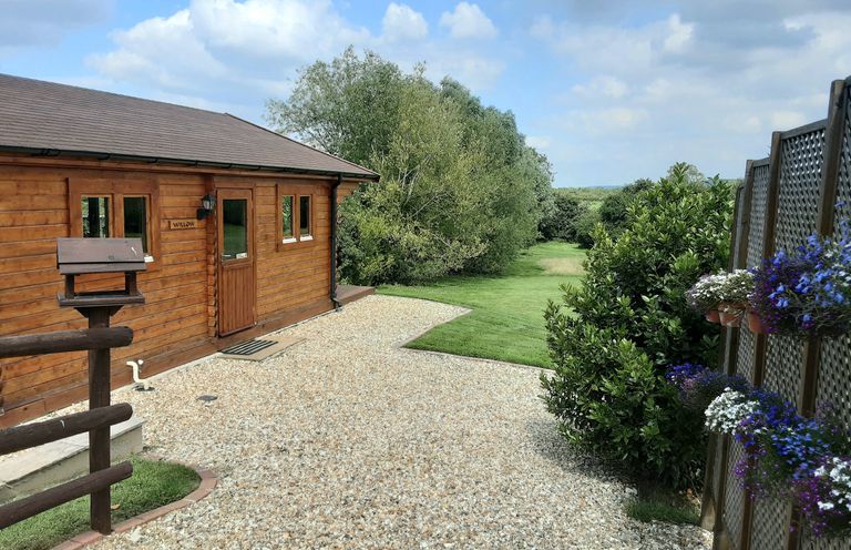 A wooden cabin with a gravel path and green lawn surrounded by trees and flowers at Pennylands Willow Lodge in Broadway