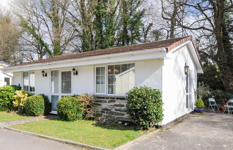 A single-story white house with shrubbery in front and folding chairs on the side at No 1 The Heathers Rosecraddoc Lodge near Liskeard