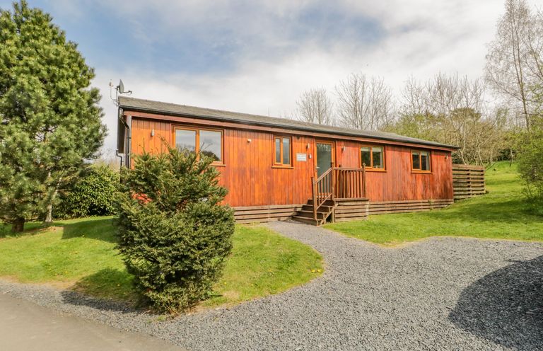 An exterior view of a wooden lodge with steps leading to the entrance surrounded by grass and trees at Cartmel Lodge in Allithwaite near Grange-over-Sands