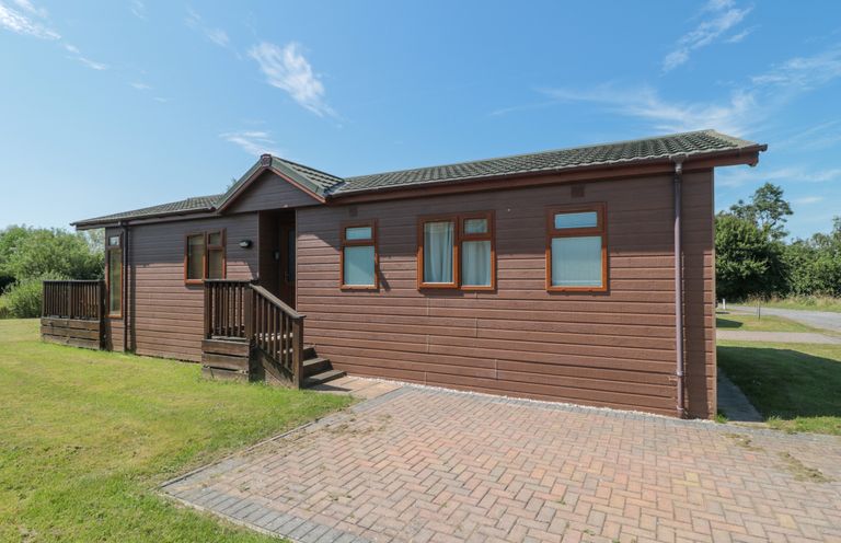 A single story wooden lodge with steps and railing at the entrance and a driveway at Harvester Lodge in Hewish near Weston-super-Mare