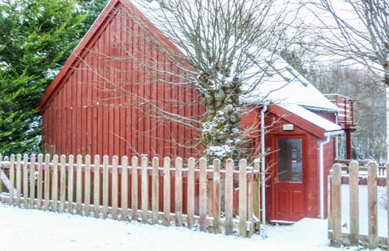 A red wooden house with a picket fence and snow on the ground and roof at Strathavon Chalet in Aviemore