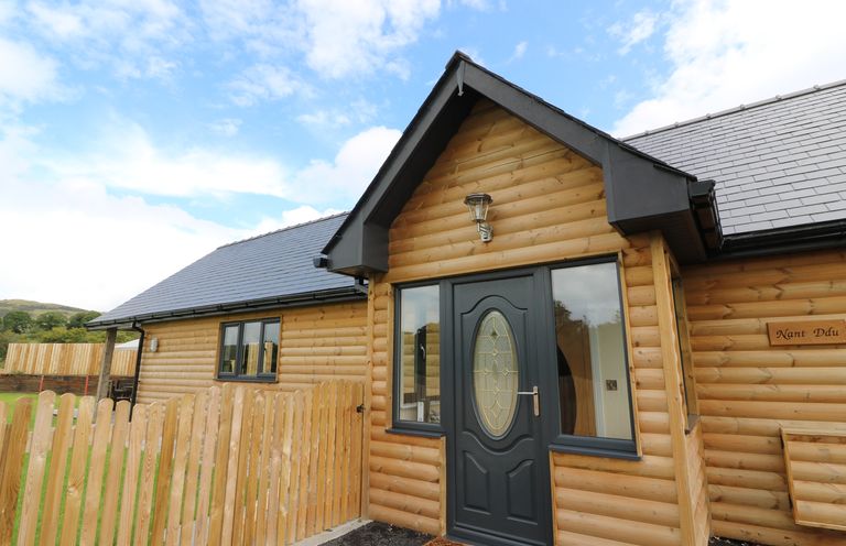 The exterior entrance of a wooden cabin with a black door and windows and a wooden picket fence at Caban Nant Ddu in Rhayader