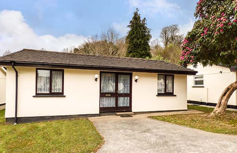 A bungalow with a driveway and grass lawn under a tree with pink flowers at Chy Lowenna Rosecraddoc Holiday Village near Liskeard