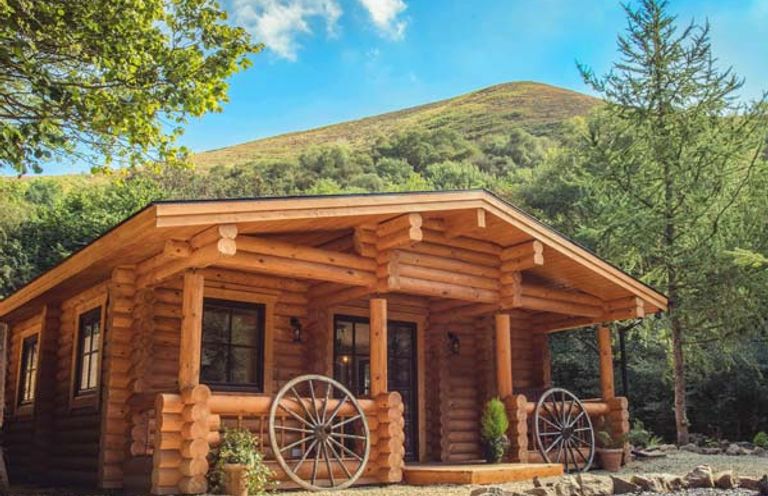 A wooden log cabin with wagon wheels on the porch and trees around at Wilderness Lodge in Stiperstones