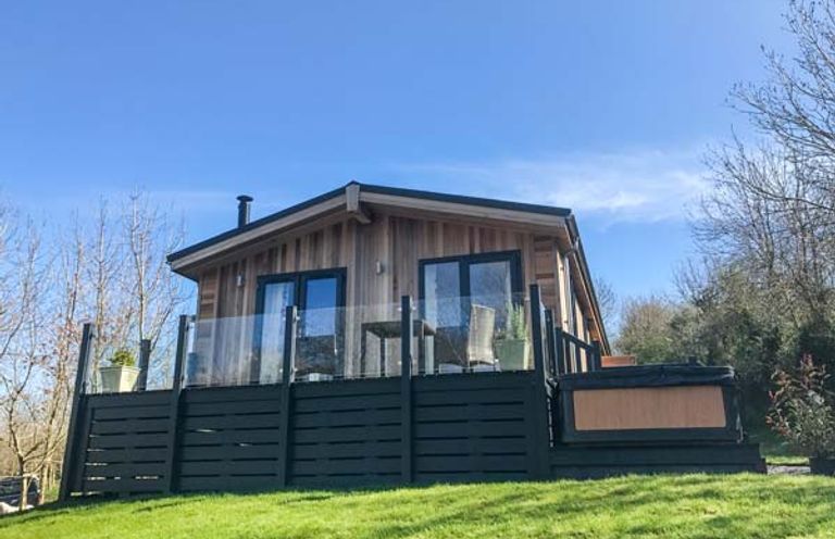A wooden cabin with glass railing on a deck and a hot tub on the side surrounded by grass at Crook Lodge in Allithwaite near Grange-over-Sands