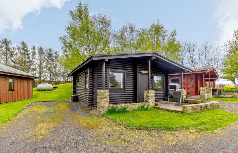 A log cabin with a porch and outdoor furniture surrounded by grass and trees at Harbottle Lodge in Longframlington near Alnwick
