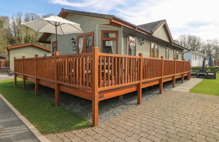 A wooden deck with railing and a white patio umbrella outside a single story cabin at Heddwch Wisemans Bridge near Saundersfoot