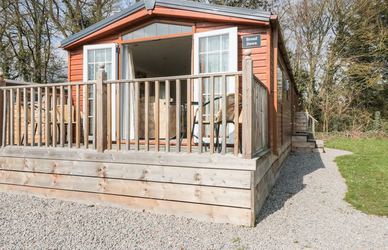 A wooden cabin with an open door and a deck with chairs surrounded by gravel and trees at Broad Down in Berrow