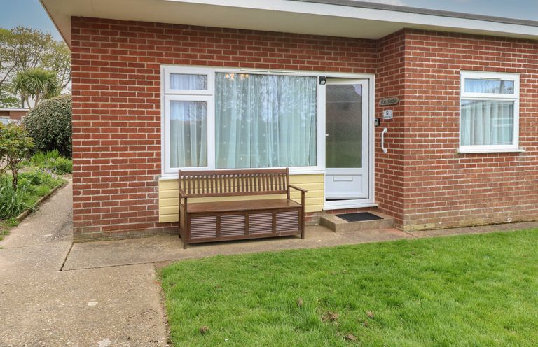 A brick house with a white door and windows a wooden bench on the concrete path and a lawn in front at En-Casa Mundesley