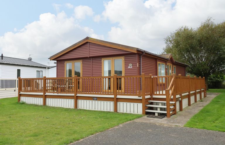 A brown single-story cabin with wooden deck and railing on a grassy plot at Jolie in Hunstanton