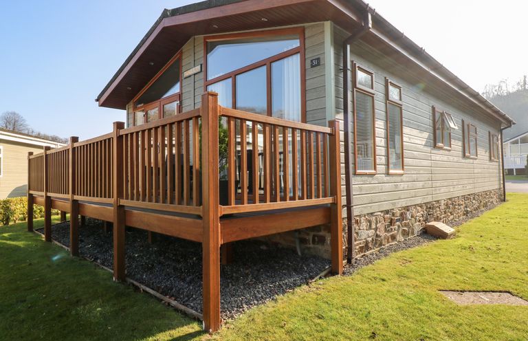 A single story house with wooden porch railing and multiple windows on a grassy area at Tranquillity - Heritage Park in Stepaside near Kilgetty