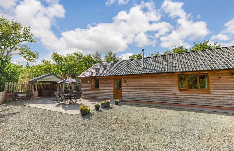 An outdoor area with a wooden building a patio table with chairs an umbrella planters and trees at Ploony Hill Lodge in Bleddfa