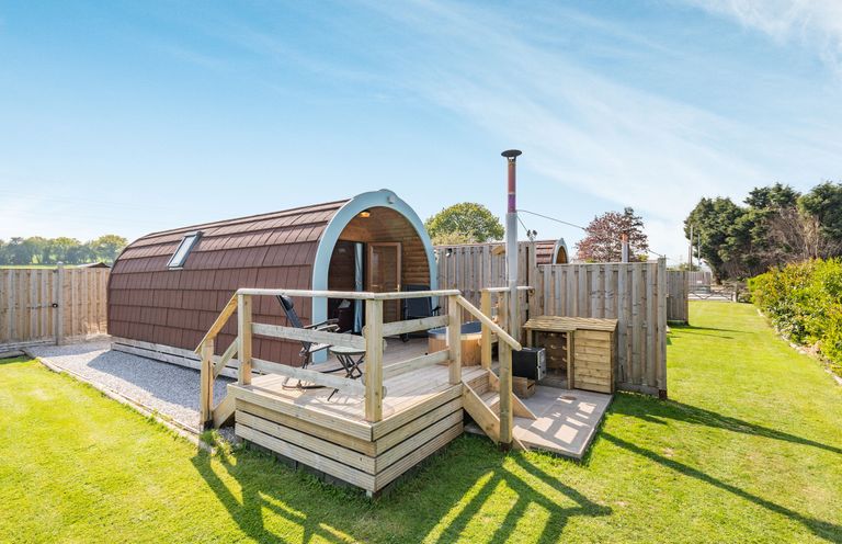 An outdoor pod accommodation with a small deck balcony and two chairs surrounded by grass at Two Hoots in Northop