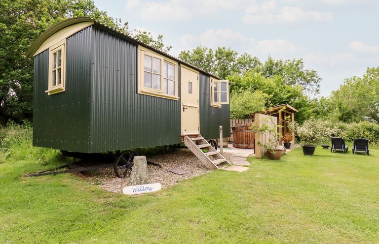 A green caravan with steps leading to the door on grass with outdoor seating and a wooden shelter at Willow Burlawn near Wadebridge