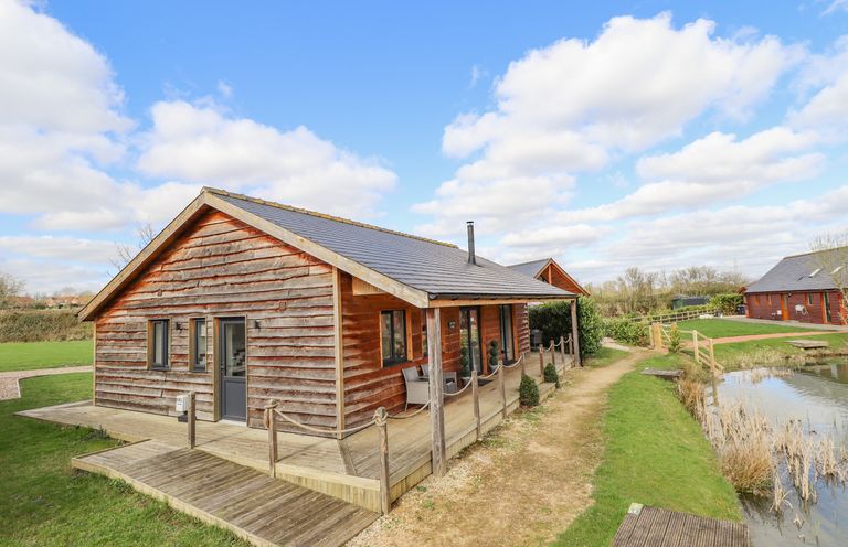 A wooden lodge with a porch next to a small pond and a gravel path at Lily-pad Lodge in Thorpe-on-the-Hill