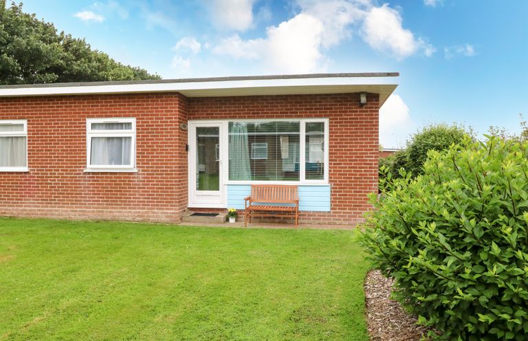 A brick house with a white door and windows a wooden bench a green lawn and bushes at 20 Siesta Mar in Mundesley