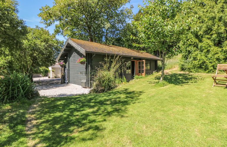 A wooden cabin with open doors surrounded by grass and trees with a chair in the garden at Pond Cabin in Stithians