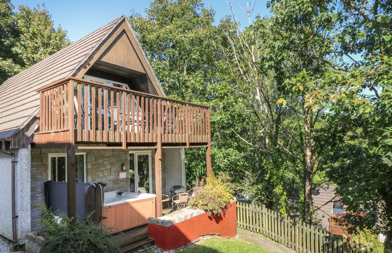 A two-story cabin with a wooden balcony and outdoor hot tub surrounded by trees at Valley Lodge 58 in Callington