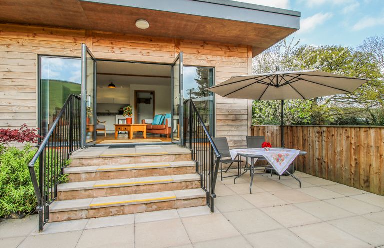 A patio with table chairs and umbrella next to steps leading to a room with open glass doors at No1 The Lodge Acton near Bishops Castle