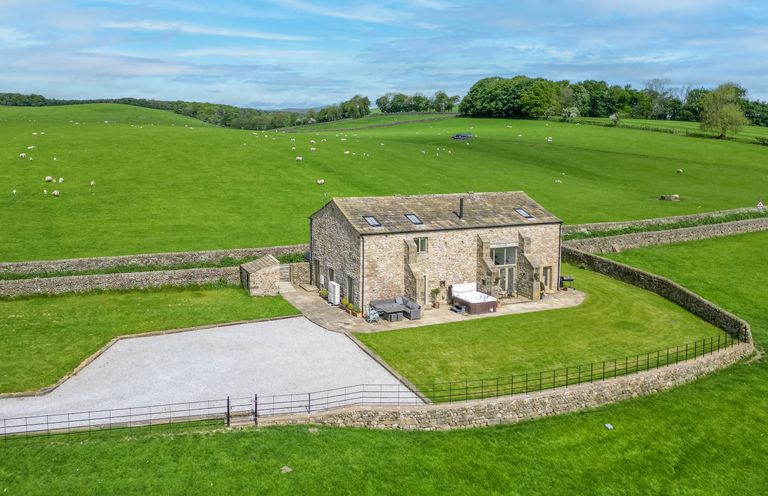 A stone house with a patio and hot tub surrounded by green fields and stone walls at Crane Field Laithe in Otterburn North Yorkshire