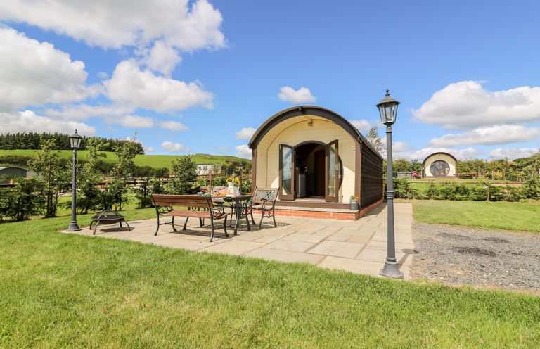 A small curved cabin with double doors open on a paved patio with a metal table and bench and two street lamps in a green field at Meadow View in Rhayader