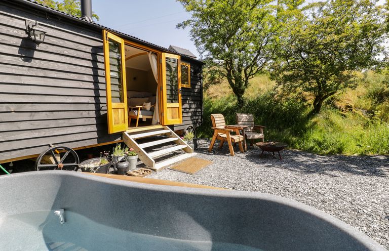 An outdoor area with a wooden hut with open doors two wooden chairs a fire pit and a small pool at Moelfre - Shepherds Hut in Llanbedr