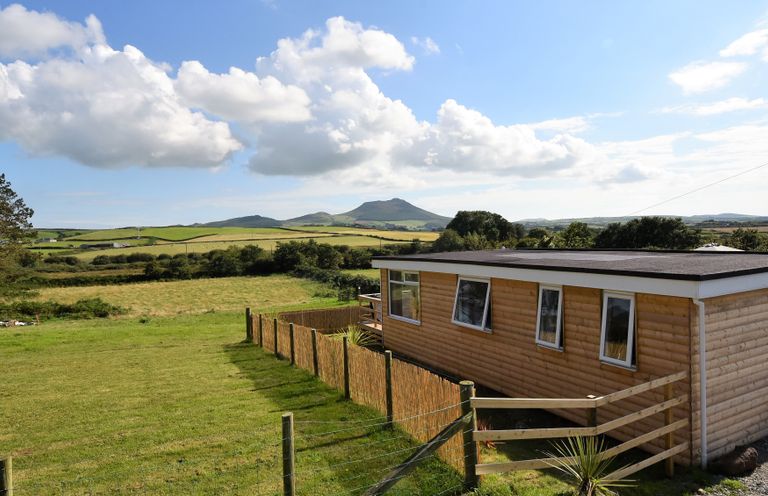 A wooden cabin next to a fenced grassy field with hills and clouds in the background at Y Caban Clyd in Morfa Nefyn