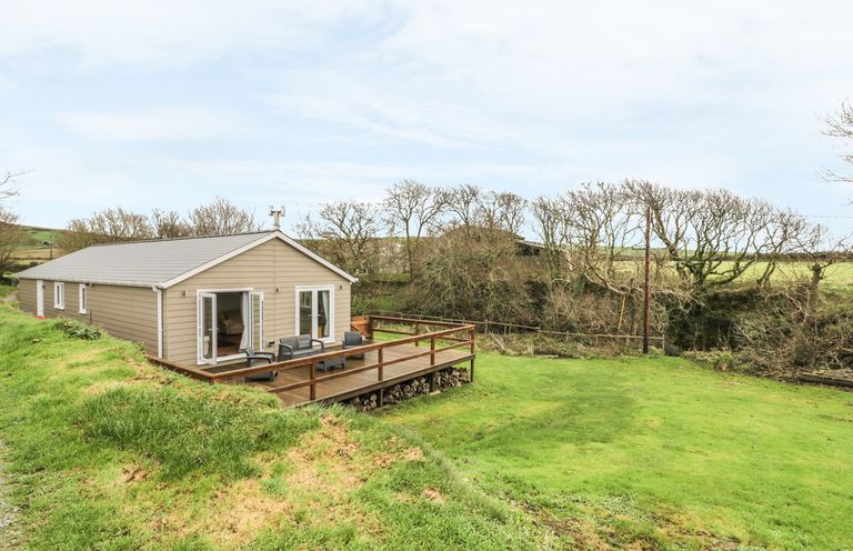 A single-story building with beige siding and a wooden deck on a grassy area with trees in the background at Nant Y Felin Lodge in Aberdaron