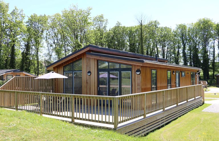A wooden cabin with large windows and a surrounding wooden deck with a table and umbrella outside in a grassy area