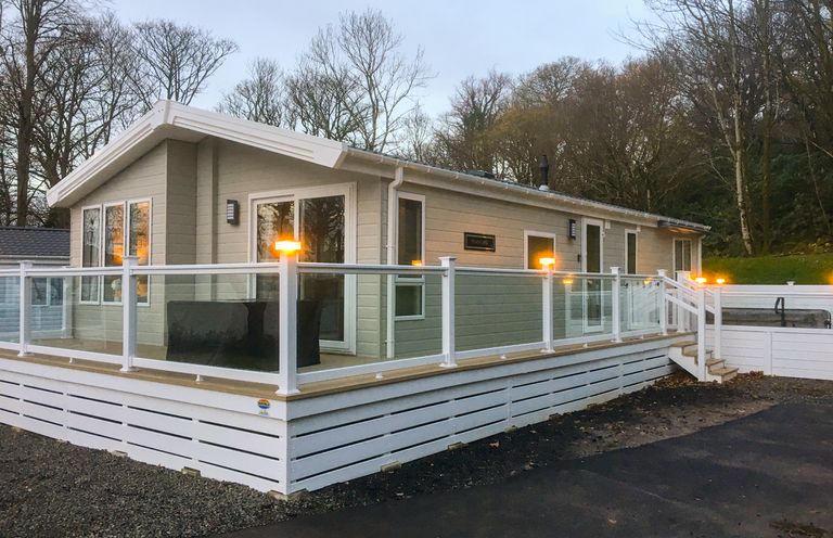 A single-story house with a glass railing deck and outdoor lighting surrounded by trees at Woodstock Lodge in Wemyss Bay
