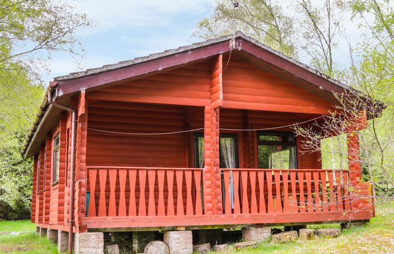 A red wooden cabin with a covered porch surrounded by trees at Otter Lodge in Strathpeffer