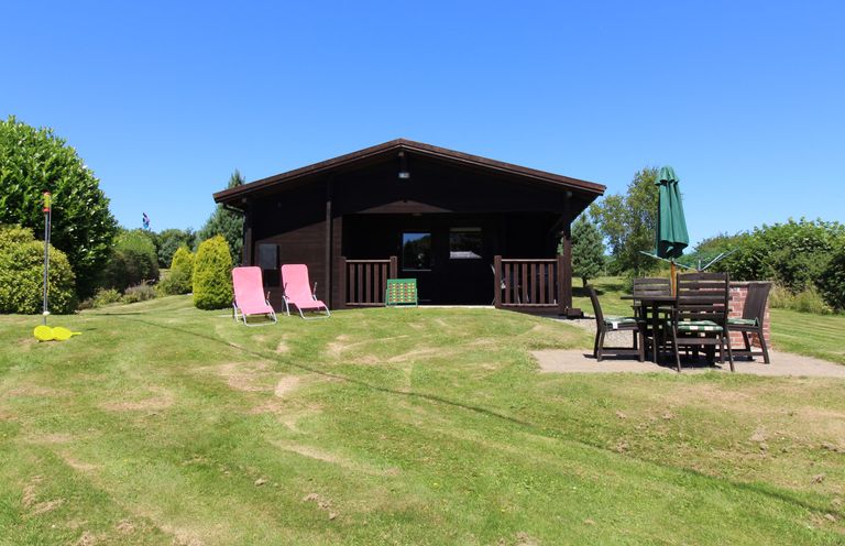 A wooden cabin with two pink chairs and outdoor dining table on a grassy lawn at Cedar Lodge in Lanivet near Bodmin