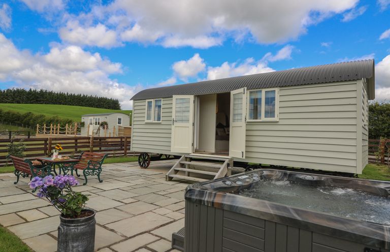 An outdoor patio area with a small shepherd's hut, hot tub, metal table and chairs with flowers on a table and a potted plant at Hillside View in Rhayader