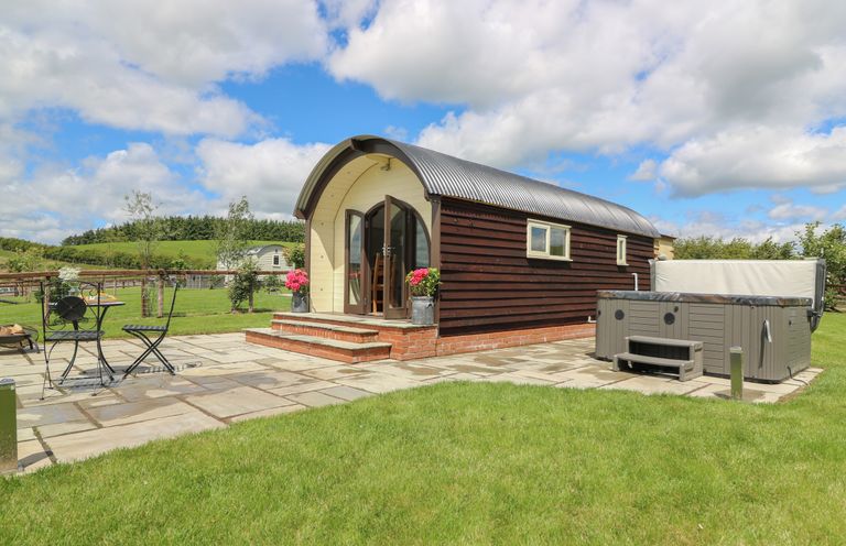 A small wooden cabin with arched roof and open double doors on a paved patio with a hot tub and metal outdoor furniture