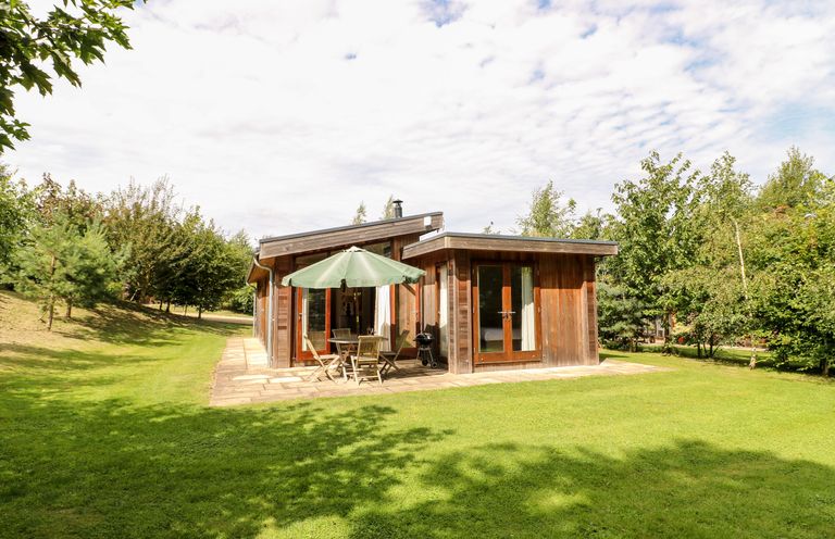A small wooden house with a patio table and umbrella on a grassy lawn surrounded by trees at Tickencote in Exton Rutland