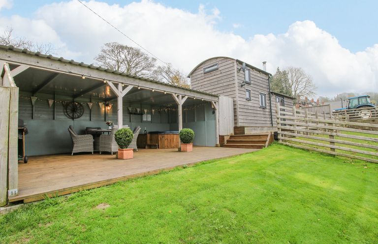 An outdoor covered patio with chairs a table and a hot tub next to a wooden cabin on a lawn at Shepherds Cabin at Titterstone in Clee Hill
