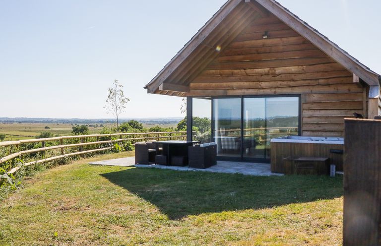A wooden cabin with large glass sliding doors and outdoor seating on a grassy area at Heron Lodge Castle Farm in Wedmore
