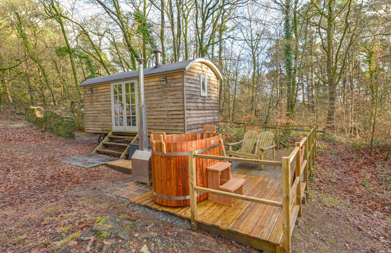 A wooden shepherds hut with a deck featuring two chairs and a wooden hot tub in a forest setting at Rock View Shepherd's Hut in Gulworthy near Tavistock