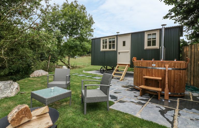 An outdoor seating area with two chairs and a table near a wooden hot tub and a small building with wheels at Orchard Burlawn near Wadebridge