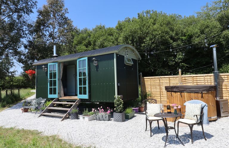 A small green shepherds hut with blue doors and outdoor seating near a hot tub at Two Moors Shepherds Hut in Hittisleigh