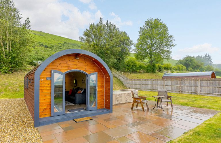 A wooden pod with open doors on a tiled patio with two chairs and a small table at Corndon Pod in White Grit near Chirbury