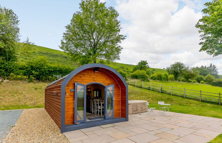 A wooden pod with glass doors open showing a dining area outside a hot tub on a stone patio at Stiperstones Pod in White Grit near Chirbury