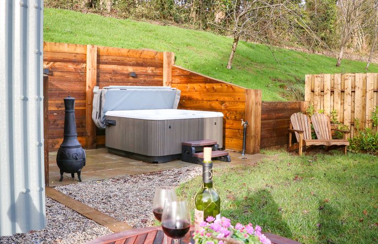 A backyard with a hot tub wooden bench a table with wine glasses and flowers and a metal chiminea at Little Silver Fox in High Bickington