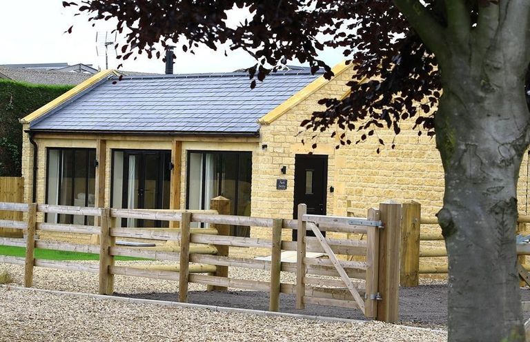 A stone building with a slate roof behind a wooden fence and a tree at Park Bothy in Wyck Rissington