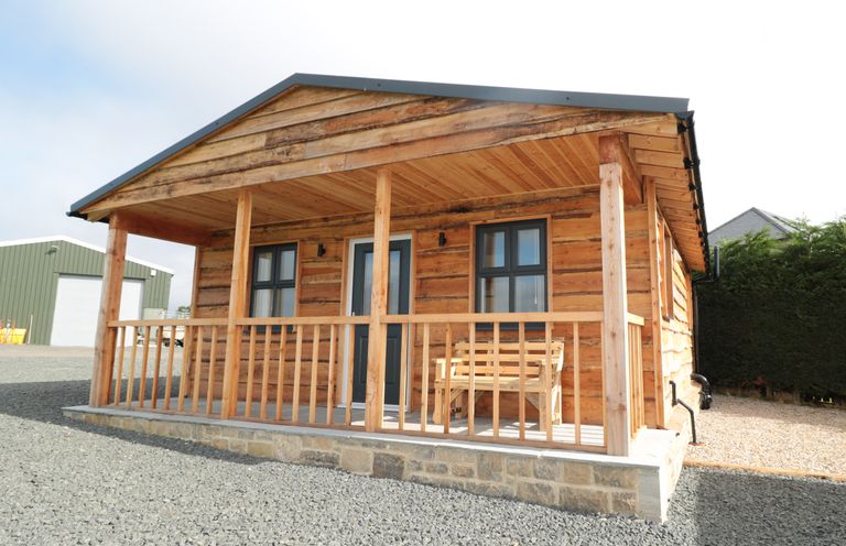 A wooden cabin with a porch and a bench outside in a gravel area at Aln in Longframlington