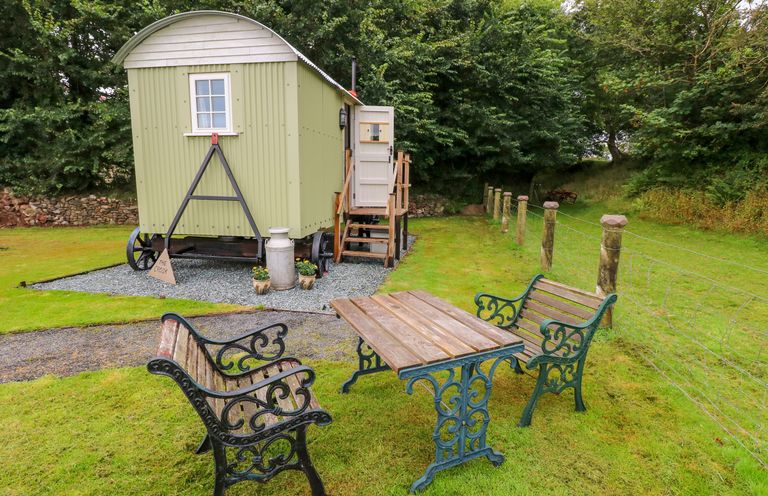 A small green shepherds hut with stairs and an open door near two wooden benches and a table on grass at Shepherds Hut - The Crook in Rosemarket near Neyland